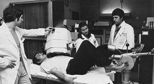 Three people in white coats operate equipment while a man lies on his back, pedalling.
