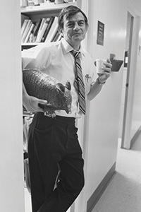 George Curry in office doorway, holding a styrofoam cup in one hand with a stuffed armadillo under his arm
