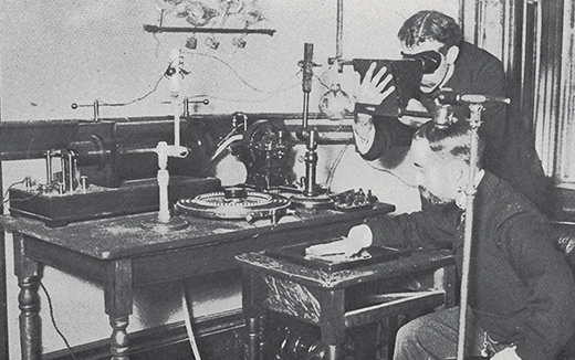 Historic photo of one man holding his hand on an X-ray plate while another operates machinery