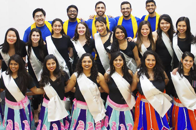 Women dancers wear black tops with pink and blue skirts and white sashes and men wear blue shirts with yellow sashes.
