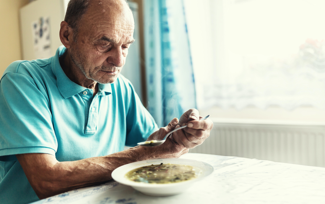 Old man with a Parkinson disease holds his arm with a hand, trying to eat a soup. 
