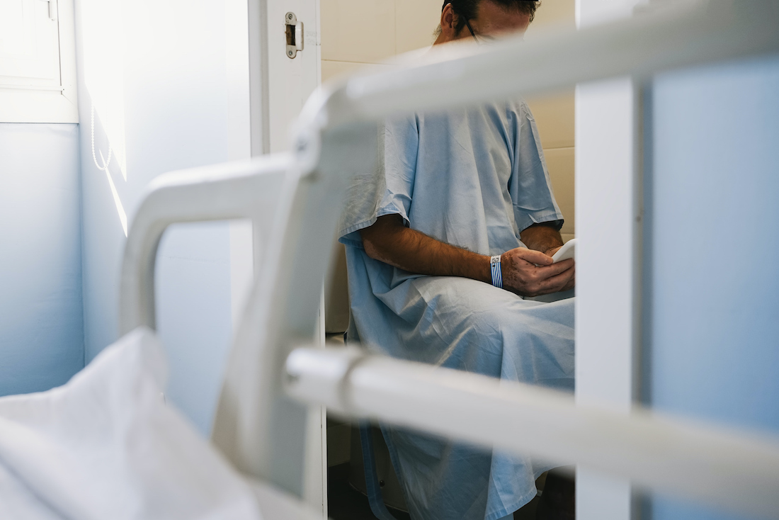 Out-of-focus close-up of the barrier of a hospital bed, with the patient sitting with his cell phone in the background