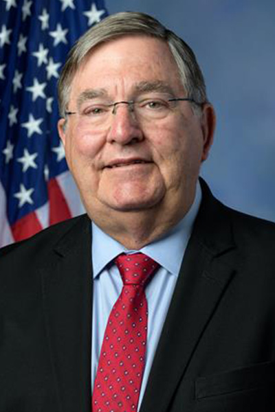 headshot of  Dr. Michael Burgess grey haired man in black blazer and red tie with U.S. flag to the left in background