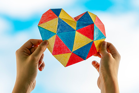 hands holding a multicolor paper heart against a blue sky, fluffy cloud background