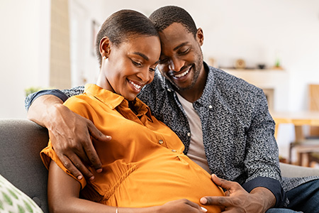 Photo of male with his arm around a female, sitting on a couch and smiling while looking at the female's baby bump.
