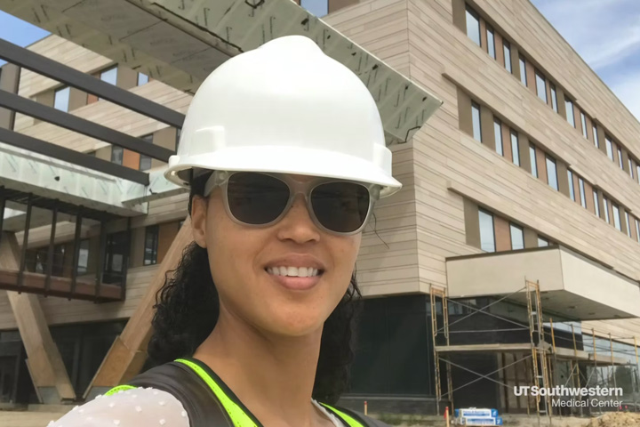 A woman wearing glasses and construction hat, standing in front of building