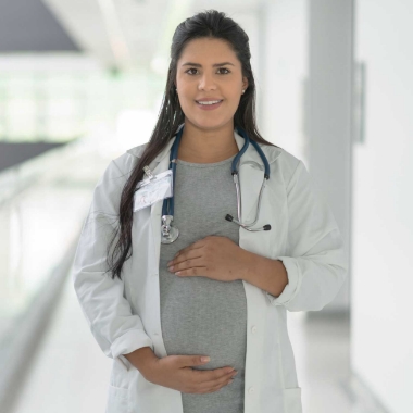 Young female physician in white lab coat smiling at camera and holding hands over her pregnant belly