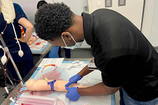 Young black man wearing blue gloves and a black collared shirt is practicing on a fake arm.