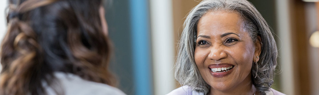 smiling black woman with gray hair and stethoscope around her neck talks to woman with brown hair in foreground
