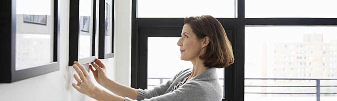 woman with brown hair and gray sweater adjusts a black picture frame on the wall to the left