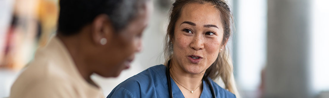 Asian female healthcare worker in blue scrubs sits and talks to older black female patient in tan shirt