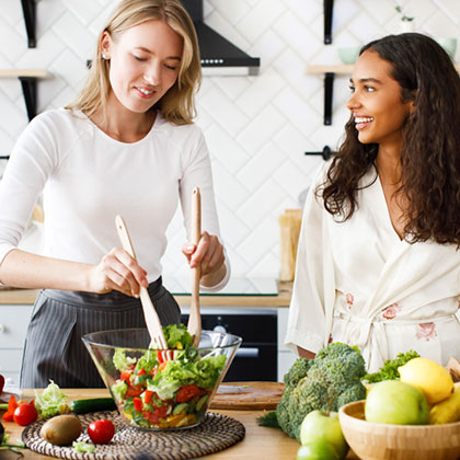 two women smile while mixing salad in kitchen