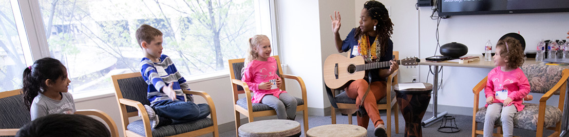 A woman sits in a chair and plays guitar as three young girls and a young boy sit in chairs around her