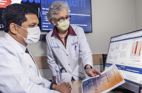 A female and male physician wearing white coats and face masks look over data on a computer monitor