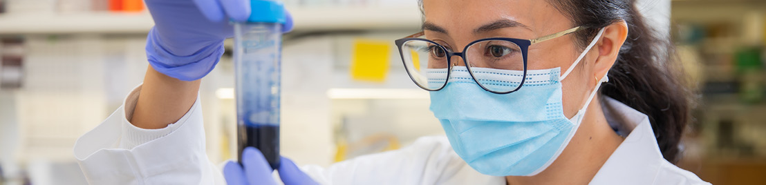 A woman wearing glasses and a blue mask looks at a test tube that contains dark liquid