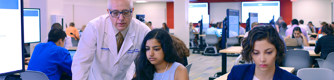 A professor in a white coat assists a female student in a classroom