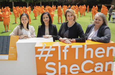 Four women sit at a table in front of orange statues of women on a lawn