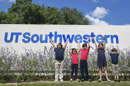 Five young children celebrate with their arms over their heads in front of a UT Southwestern sign