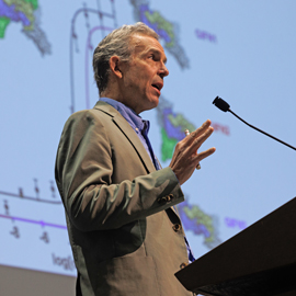 A man with grey hair standing at a podium, speaking at a conference 