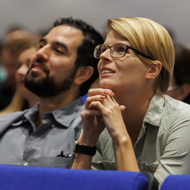A woman with blonde hair, wearing glasses, sitting next to a man with dark hair, looking up at conference
