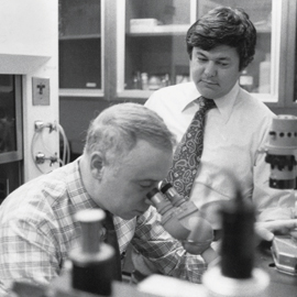 Two men in a black and white photo examining particle under microscope in a lab 