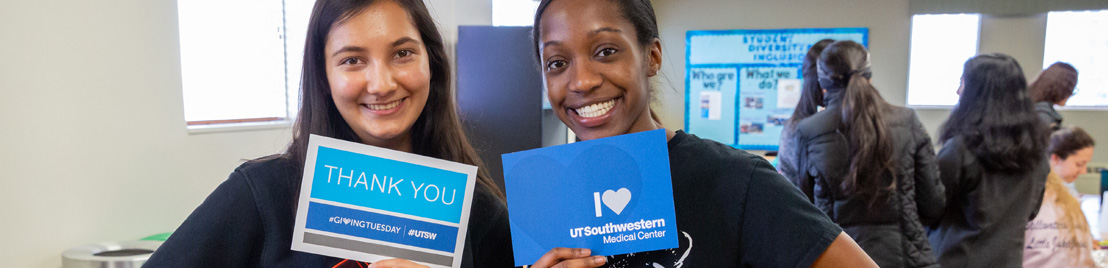 Two woman holding signs that say I love UTSW and #GivingTuesday 