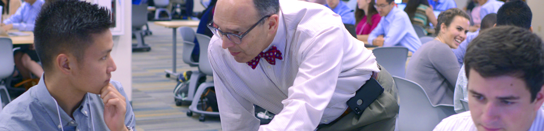 A professor with a white shirt and red bowtie, assisting a male student in a classroom