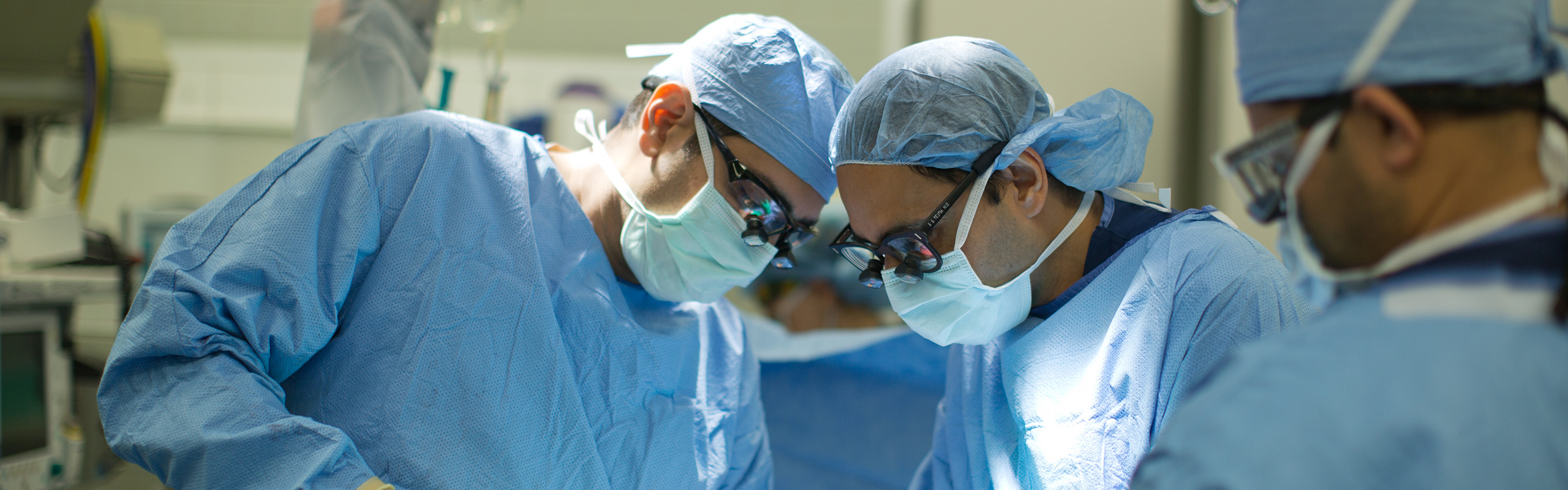 Doctors and nurses in scrubs and masks bending over an unseen patient performing Plastic Surgery.