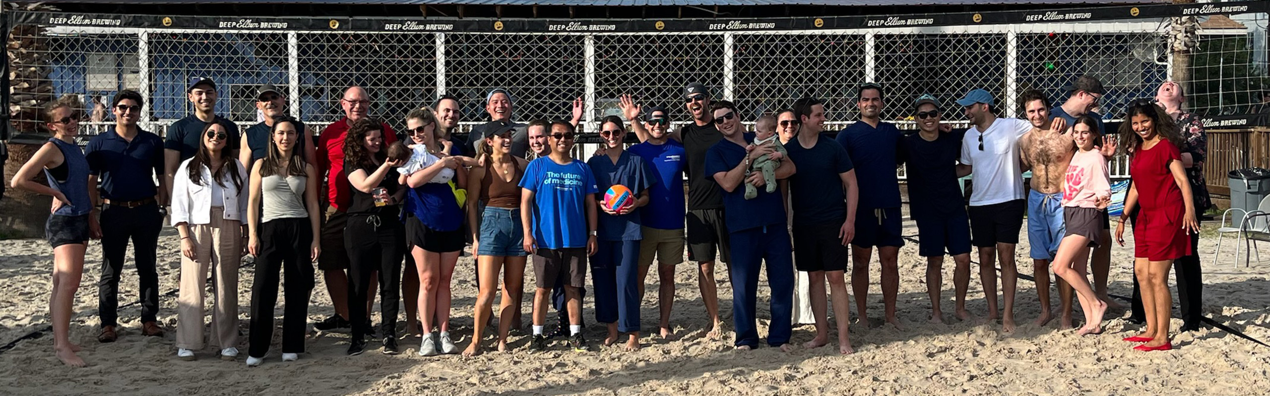 The Plastic Surgery team and families on on outing. The group is dressed casually, standing on sand in front of a fence.