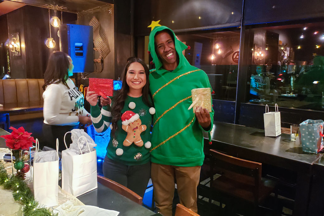 A woman wears a gingerbread man sweater and a man wears a sweater that has a hood that makes the sweater shaped like a Christmas tree