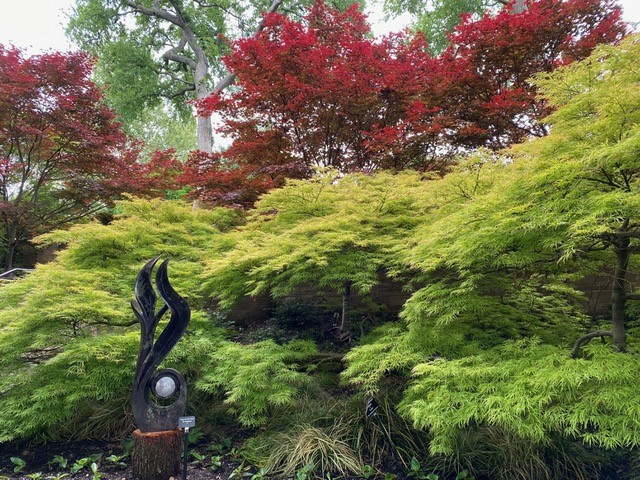 graceful metal sculpture in front of thick trees with vibrant green and red foliage