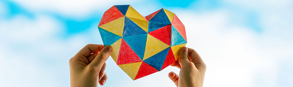 Photo of multi-colored paper heart being held up against a blue sky with clouds