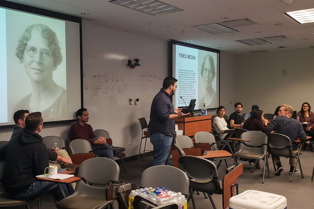 A male student stands in front of a screen with a photo of Ynez Mexia, a Mexican-American botanist