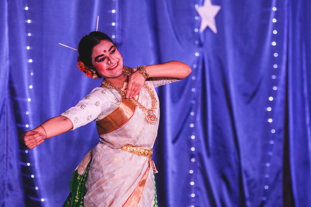 A woman dances with a red dot on her forehead and while wearing a white dress with a gold sash and white sticks in her hair