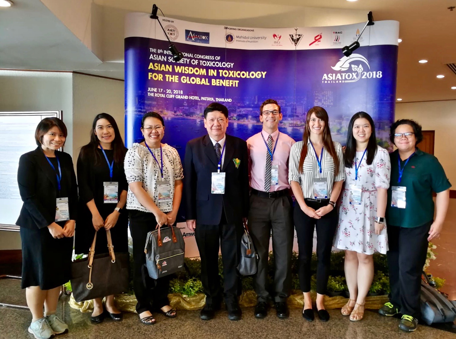 Group photo of Christian Davidson, Taylore King, and the Ramathibodi Poison Center toxicology team in front of the AsiaTox Conference sign