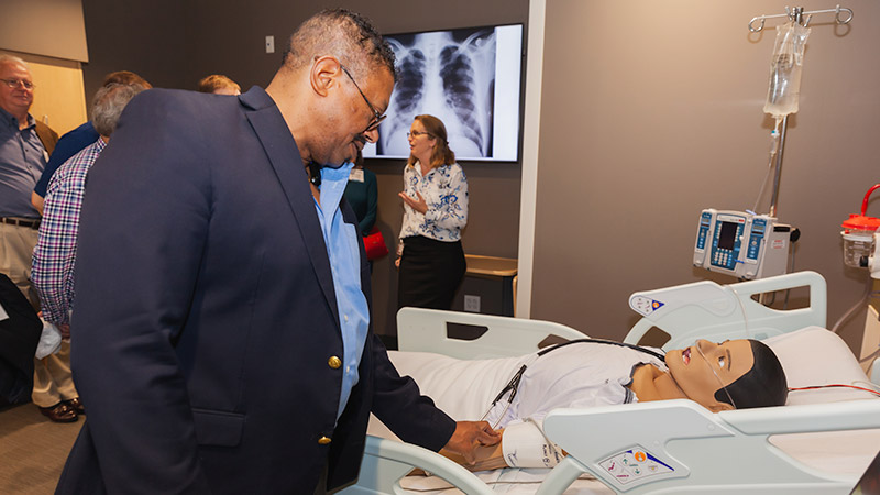 Man in blazer standing next to a hospital bed with a patient dummy laying in it.