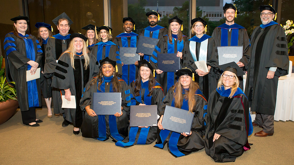 Group of graduates wearing black robes and blue sashes, holding their diplomas or programs.