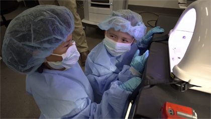 Two children dressed in operating scrubs observe a machine at Sim Center.