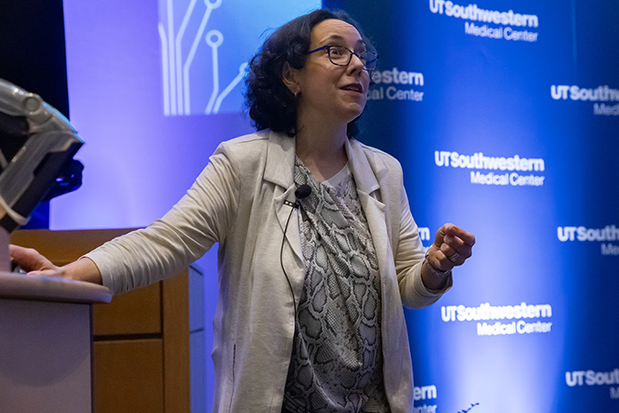 dark haired woman with glasses wearing tan jacket speaks next to podium on stage