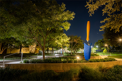 Scultpure of a trowel with a blue blade and sand colored handle, positioned amongst greenery at night