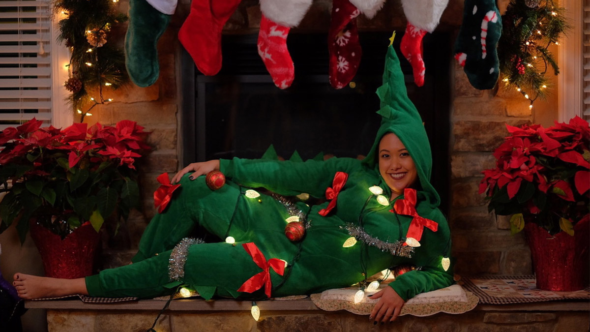 Woman dressed in green onesie covered in bows and lights reclining on a fireplace