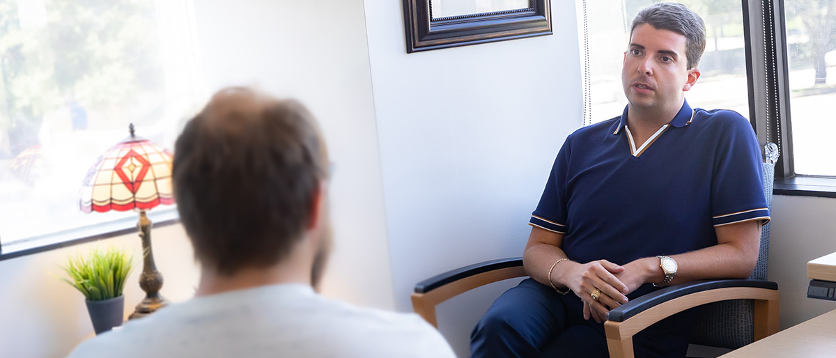 Dark haired man wearing a blue shirt and slacks, seated, talking with another man who's back is to the camera.