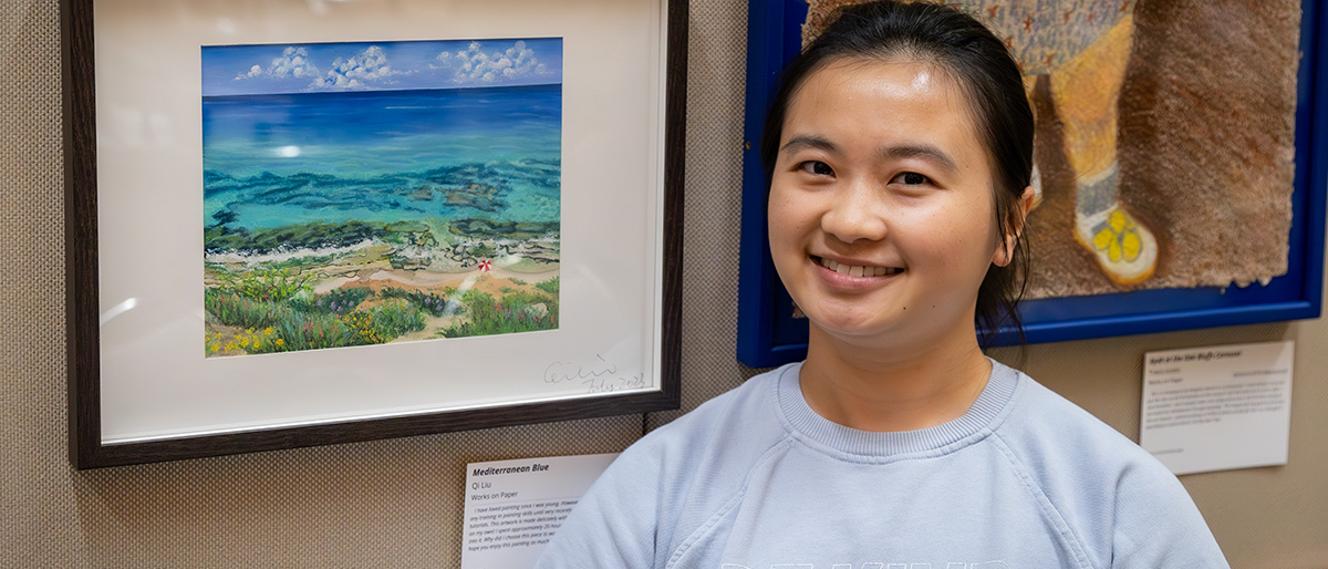 Smiling woman with dark hair standing next to the painting of the the ocean and beach she entered in the art show.