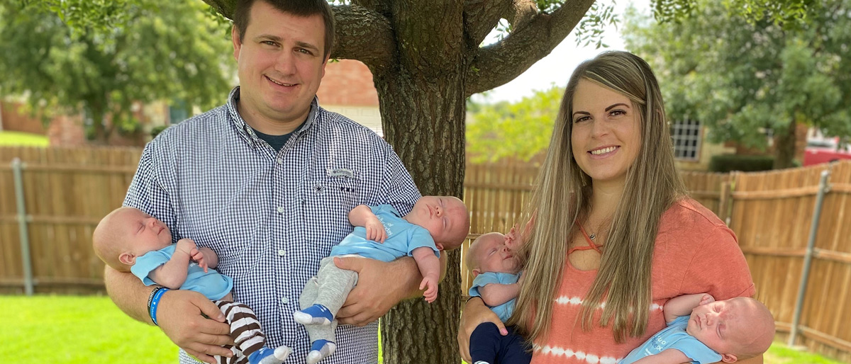 Man and woman standing in yard holding two babies each, with a young boy standing between them