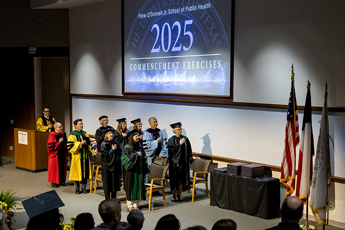 Speakers on stage at the Peter O'Donnell Jr. School of Public Health 2025, Commencement Exercises face the flags with right hands over their hearts.