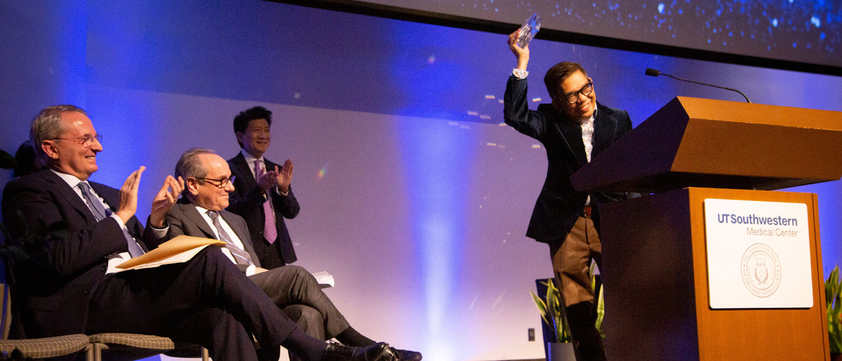 Man holding glass award over his head, smiling, while people on stage applaud