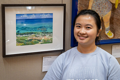 Smiling woman with dark hair standing next to the painting of the the ocean and beach she entered in the art show.