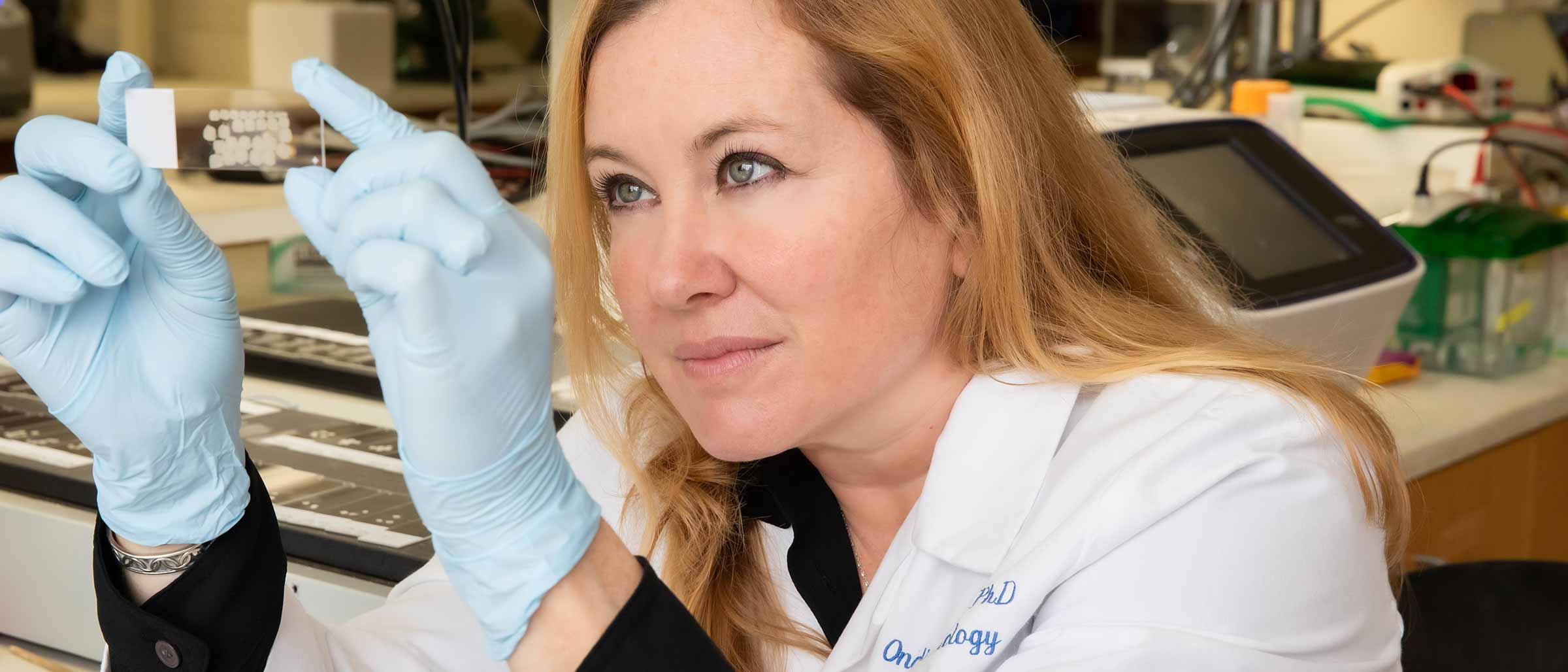 Woman with golden brown hair, wearing white lab coat and pale blue gloves, looking at slide