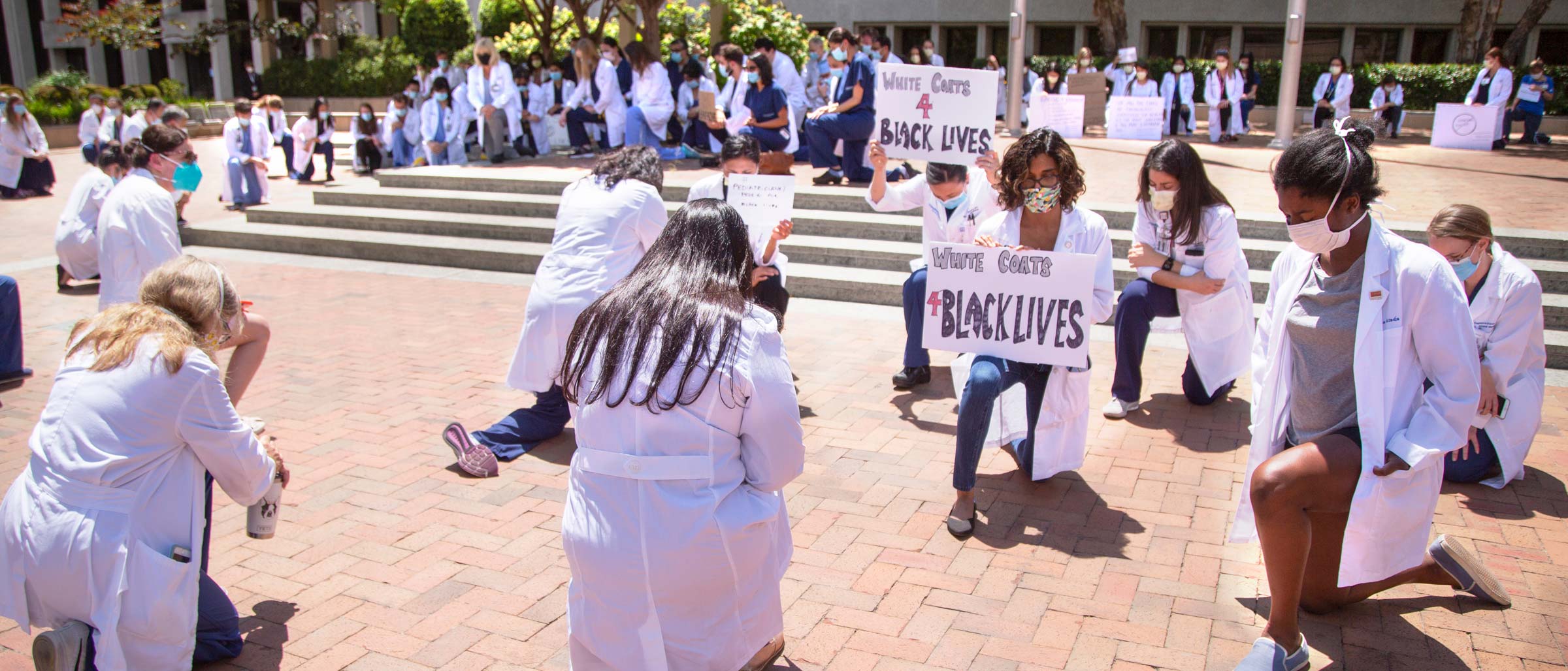 Crowd of people kneeling, wearing white lab coats and holding signs
