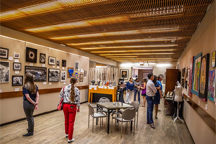gallery room with table and 4 chairs in center, groups of people mingling and looking at arwork on walls and in display cases